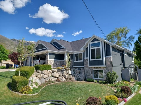 Modern two-story house with stone and white trim, solar panels on roof, surrounded by landscaped yard with boulders and manicured shrubs