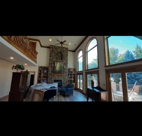 Spacious modern living room with soaring ceilings, stone fireplace, large windows overlooking trees, and wooden balcony railing