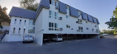 Modern multi-story residential building with white siding, dark roof, and ground-level parking garage with vehicles